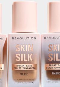 Tre flasker med Revolution Skin Silk Radiant Satin Serum Foundation. Flaskene har rosa lokk og viser skyggenumrene: F12.7NC og F14.9NC.