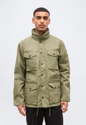 Mężczyzna ubrany w oliwkowo-zieloną kurtkę z czterema kieszeniami z patkami zapinanymi na guziki oraz wysokim kołnierzem, stojący na jednolitym jasnym tle.