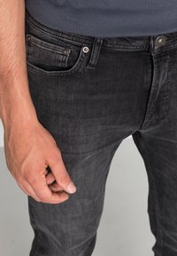 Jeans de mezclilla negros de corte ajustado con textura suave, diseño de cinco bolsillos y detalles de botón y remaches de metal. Mano colocada cerca del bolsillo.