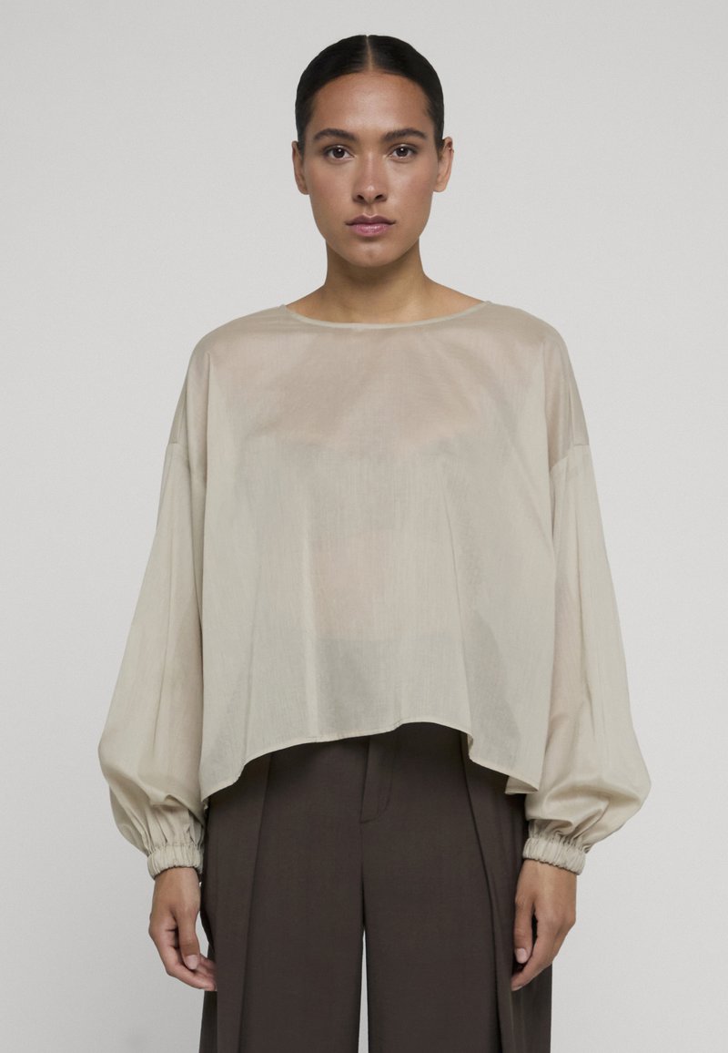 Femme aux cheveux foncés tirés en arrière, portant un chemisier ample beige clair à manches longues avec des poignets froncés et un pantalon large marron foncé.