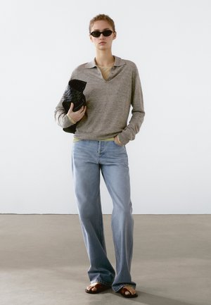 Pull en gris clair à manches longues avec col, superposé sur une chemise verte, associé à un jean bleu clair et des sandales marron. Tenant une pochette noire.