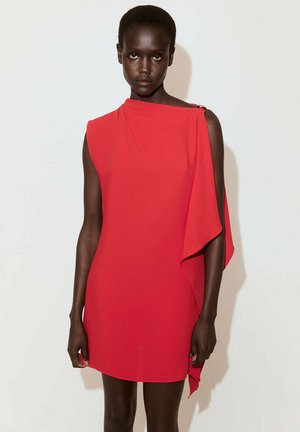 Femme debout dans une robe rouge sans manches avec un détail de cape asymétrique, sur un fond uni clair.