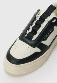 Valge ja must sneaker, millel on tekstureeritud nahast pealis, pehmendatud keel, elastsed paelad ja paks, tekstureeritud kummitald. Brändi logo küljes.