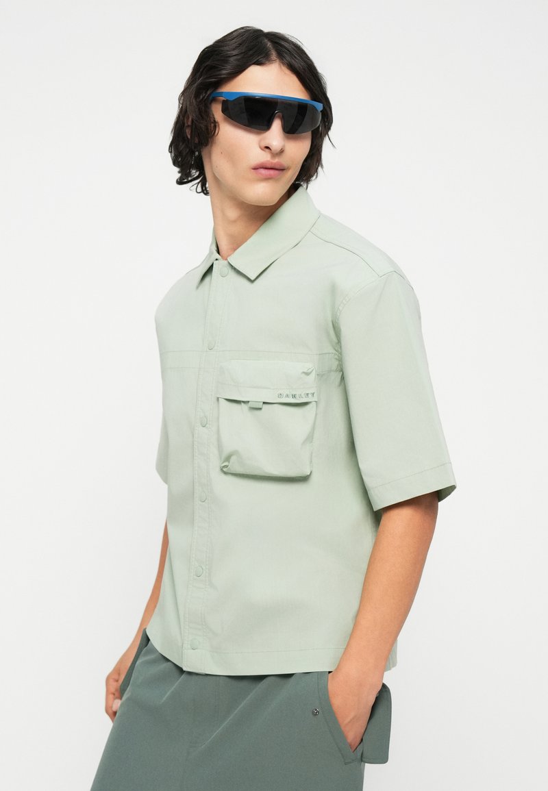 Camisa de manga corta, de color verde claro y hecha de una tela suave, con un bolsillo en el pecho y cierre de botones. Llevada con gafas de sol oscuras.
