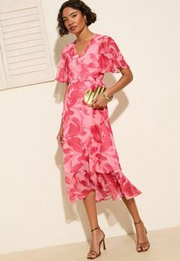 Robe rose avec un motif floral, manches courtes à volants et un design croisé. Associée à des talons à lanières et un sac à main doré.