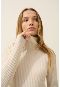 Béžový žebrovaný svetr s vysokým límcem a dlouhými rukávy, který se vyznačuje jemnou texturou a přiléhavým střihem, zdůrazňující vertikální pruhy.