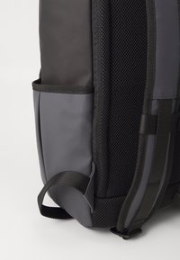 Gray backpack featuring a smooth fabric side, breathable mesh panel, and side pocket. Straps and stitching in contrasting black.