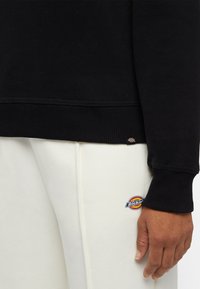 Zwarte sweatshirt met geribbelde boorden en zoom, gecombineerd met crèmekleurige joggingsbroek met een ronde Dickies logo patch. Zachte textuur.