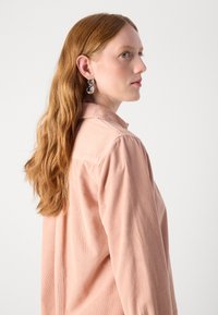 Light pink corduroy shirt with collar, visible textured fabric, worn with silver statement earrings, long wavy hair on one side.
