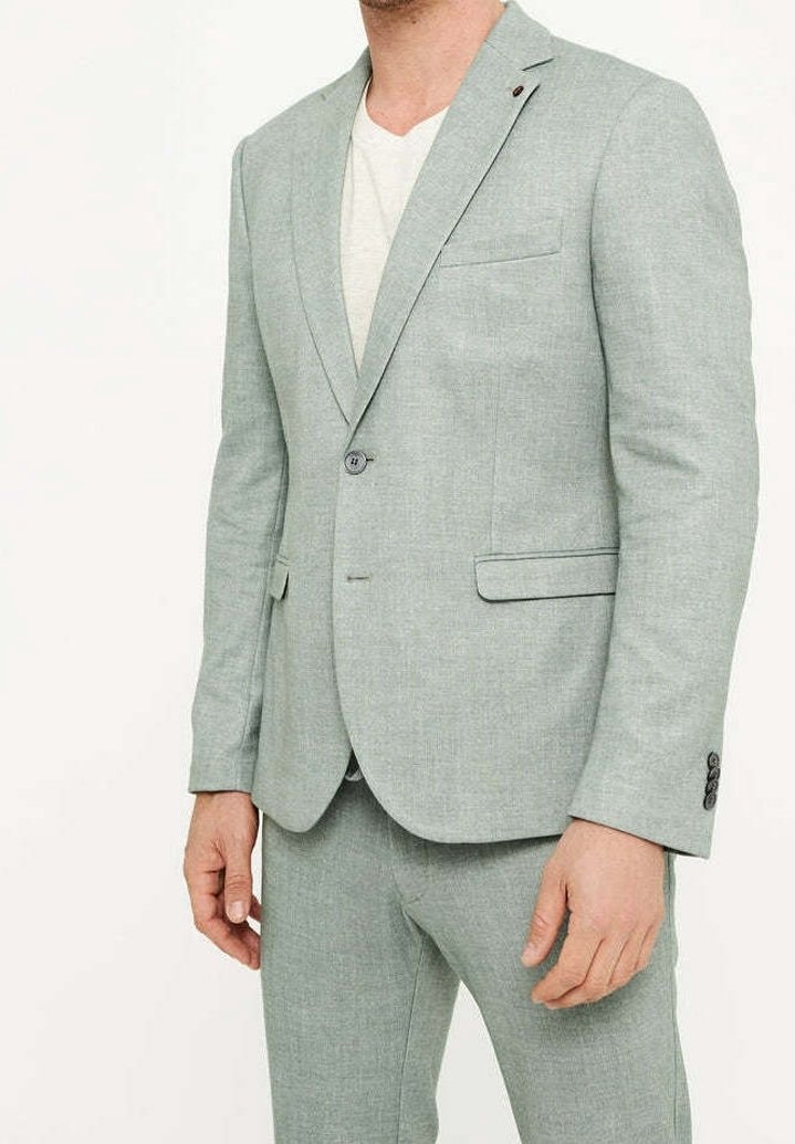 Homme portant une veste de costume gris clair et un pantalon assorti sur un t-shirt blanc, debout devant un fond clair uni.