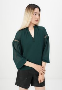 Kvinne med langt ombre-hår iført en mørkegrønn bluse med blonder detaljer på ermene og svarte skinnshorts, poserer sidelengs.