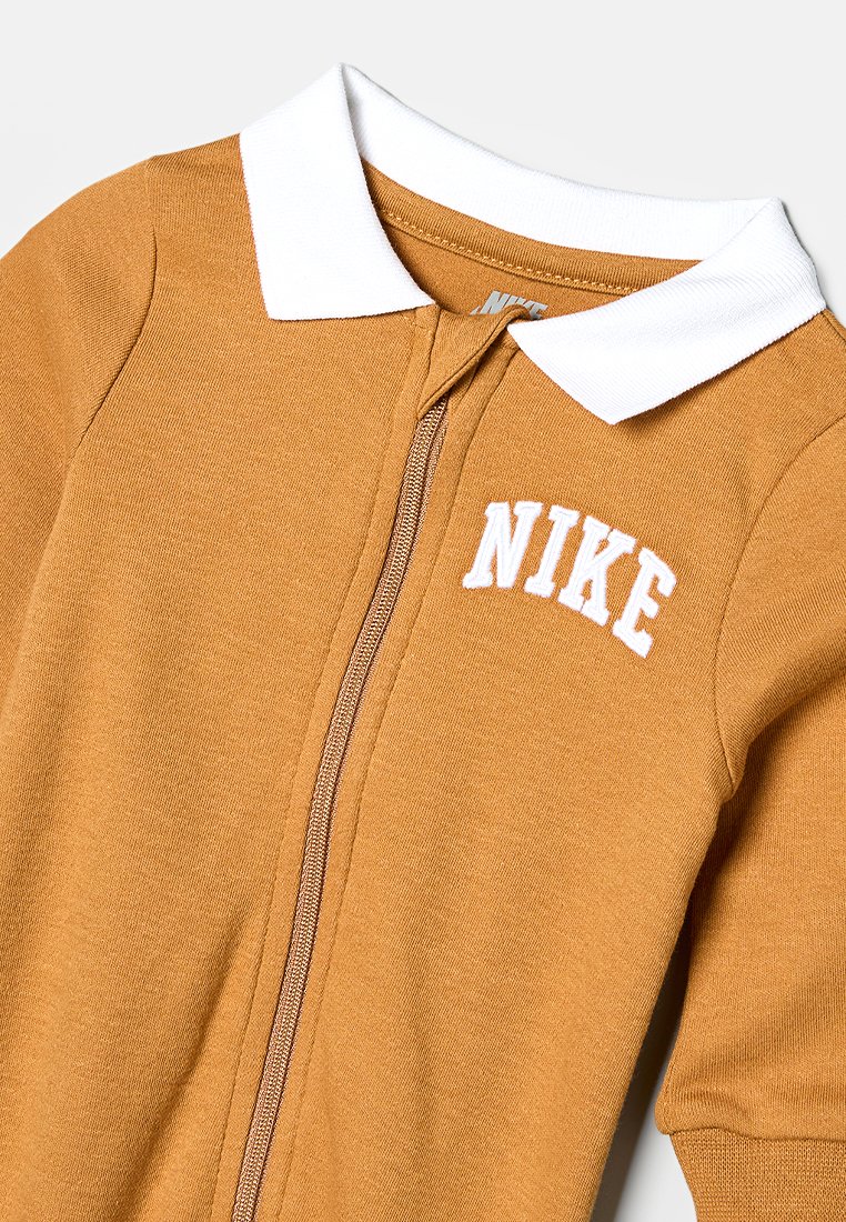 Prenda con cremallera de Nike en tejido amarillo mostaza. Presenta un collar blanco, un logo bordado y una cremallera suave. Diseño atlético y suave.