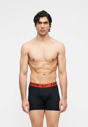 Homme torse nu debout devant un fond uni, portant un boxer Nike noir avec une ceinture rouge, faisant face à l'avant avec une expression neutre.