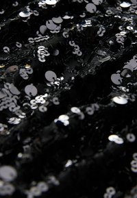 Svart tyg täckt med glänsande, reflekterande silverpaljetter som skapar en strukturerad och mönstrad yta med varierande storlekar av paljetterna.