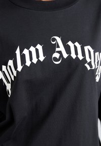 Juodas marškinėliai su balta gotikine užrašu "Palm Angels", kuris yra įspaustas ant krūtinės išlenktoje arkos formoje.