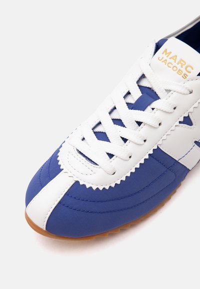 Gros plan d'une basket bleue et blanche avec des lacets blancs et "Marc Jacobs" en doré sur la languette, semelle en caoutchouc marron visible en bas.