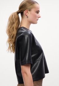 Top em couro preto com mangas curtas; textura suave e corte solto. Detalhes de costura notáveis na parte da frente. Modelo combinado com uma saia estampada.