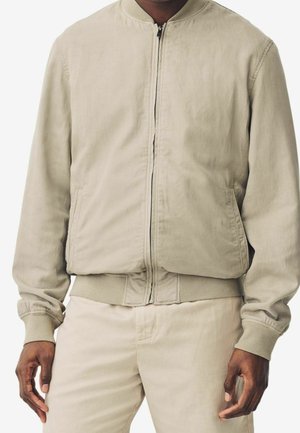 Homme portant une veste bomber beige avec zip sur le devant et un pantalon beige assorti, debout devant un fond blanc.