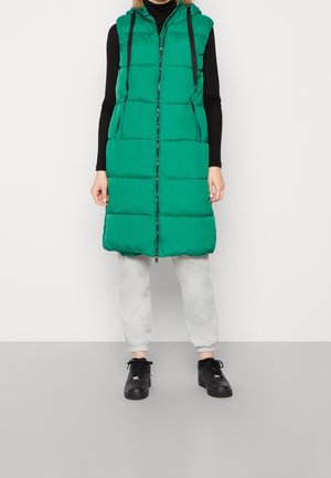 Osoba ubrana w zieloną pikowaną kamizelkę z kapturem sięgającą do kolan, czarną bluzkę z długim rękawem, jasnoszare spodnie dresowe i czarne trampki, stojąca na białym tle.