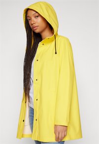 Yellow raincoat with a hood, featuring a snap button closure, side pockets, and a relaxed fit. The material appears waterproof and smooth.