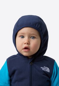 Vestă bleumarin din fleece cu glugă, mâneci albastre, fermoar frontal și logo alb. Textură netedă, cu o prindere strânsă în jurul feței.