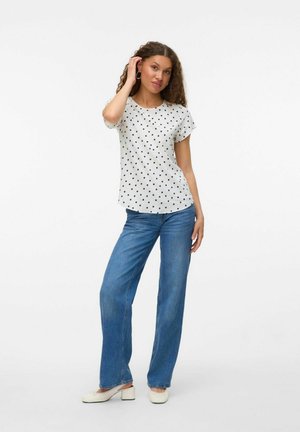 Witte top met korte mouwen en zwarte stippen, gecombineerd met wijde blauwe jeans en witte schoenen. Krullend haar en een casual pose.