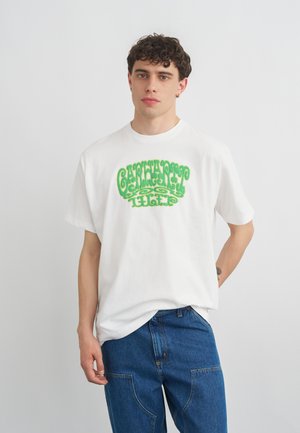 Junger Mann mit lockigem Haar trägt ein weißes T-Shirt mit grünem Grafiktext und lockeren blauen Jeans, steht vor einem schlichten hellgrauen Hintergrund.