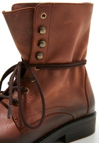 Botte en cuir marron à lacets avec œillets et crochets métalliques, semelle noire, et un lacet marron foncé noué entourant la cheville.