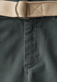 Tejido verde entrelazado con un sutil patrón de espiga, que presenta un cinturón de tela beige asegurado con un anillo metálico.