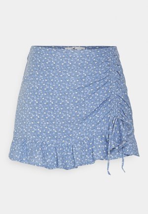 Mini-jupe bleue à imprimé floral blanc, détail froncé sur le côté droit et ourlet volanté sur fond blanc.