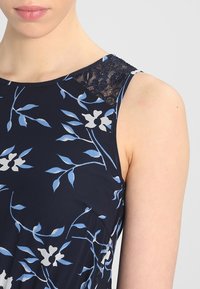 Marinblå klänning med blommönster i blått och vitt. Har spetsdetaljer vid axlarna. Tillverkad av ett mjukt, lätt material.