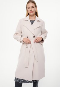 Jasnoróżowy wełniany płaszcz trenchant z podwójnym zapięciem, klapami, akcentami guzikowymi, z paskiem w talii i długimi rękawami.