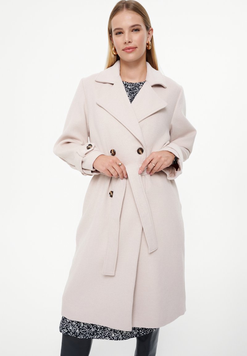 Jasnoróżowy wełniany płaszcz trenchant z podwójnym zapięciem, klapami, akcentami guzikowymi, z paskiem w talii i długimi rękawami.