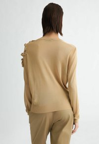 Maglione lavorato a maglia beige con dettagli arricciati testurizzati sulla spalla e polsini a costine. I materiali sono morbidi e leggeri, con una vestibilità comoda.