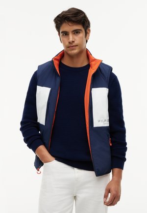 Młody mężczyzna w granatowym swetrze, granatowo-białej kamizelce z pomarańczową podszewką oraz białych spodniach, stoi z jedną ręką w kieszeni.