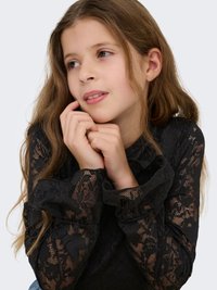 Juodas nėriniuotas ilgomis rankovėmis palaidinė su raukšlėtomis rankogaliais, puošta gėlėtu raštu ir aptempto dizaino, dėvima vaiko su ilgais plaukais.