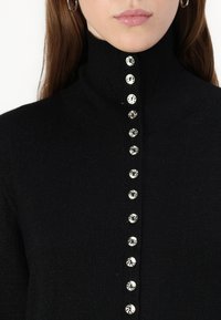 Maglione nero a maglia con colletto alto e una fila di bottoni a spirale argentati sul davanti, che mostra un tessuto testurizzato.