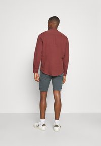Brunlanget skjorte med knapkrave, parret med grå cargo shorts. Hvide sneakers med beige accenter. Minimalistisk baggrund.