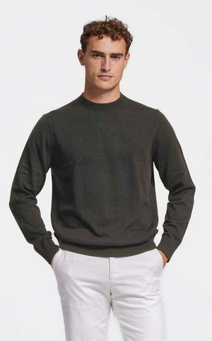 Man met krullend haar die een donkergroene trui met ronde hals en witte broek draagt, staand met handen in de zakken tegen een effen achtergrond.