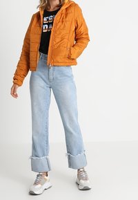 Orange puffig jacka med huva, dragkedja och texturerad yta, tillsammans med ljusblå vida jeans och klumpiga vita sneakers.