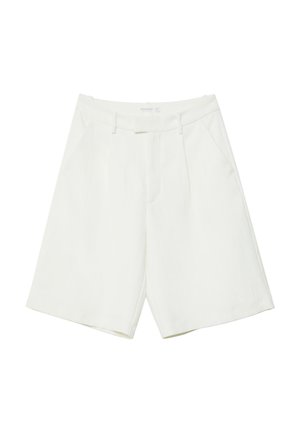 Person iført hvid kort blazer, matchende knælange shorts, hvide vævede sko og en rød vedhæng halskæde, der står på en rund brun ottoman.