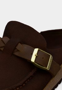 Κοντινό πλάνο σε σκούρο καφέ παπούτσι από σουέντ με ραφές και δερμάτινο λουράκι κλεισμένο με ορειχάλκινη αγκράφα.