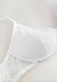 Biały koronkowy biustonosz z kwiatowym wzorem, z usztywnianymi miseczkami i regulowanymi ramiączkami, leżący na jasnej powierzchni.