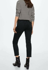 Femme portant un jean noir court, des bottines noires à talons et un t-shirt à manches longues rayé noir et blanc, debout devant un fond uni.