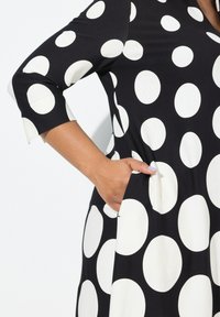 Svart klänning med vita prickar, trekvartslånga ärmar och fickor. Slät tyg, A-linjefasong, med rundade mönsterdetaljer.