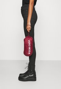 Bourgognefarvet blødt stofpose mærket "VERO MODA", holdt af en person iført sorte skinny jeans og sorte snørestøvler med plateau.