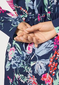 Vestido floral em azul-marinho com padrões vibrantes em rosa e verde. Apresenta mangas longas e uma cintura elástica. Mãos unidas nas costas.