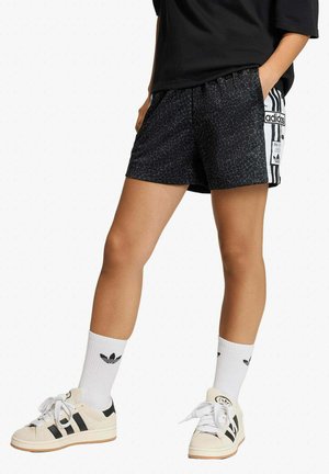 Persona che indossa pantaloncini Adidas neri con motivo a fantasia e strisce laterali, calzini bianchi Adidas al polpaccio e scarpe da ginnastica bianche Adidas con strisce nere.