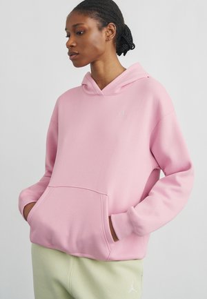 Kobieta z warkoczami, ubrana w jasnoróżową bluzę z kapturem i jasnobeżowe spodnie, z rękami w kieszeniach bluzy.