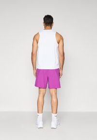 Un homme debout faisant face à un mur, vêtu d'un débardeur blanc, de shorts violets, de chaussettes blanches et de baskets blanches.